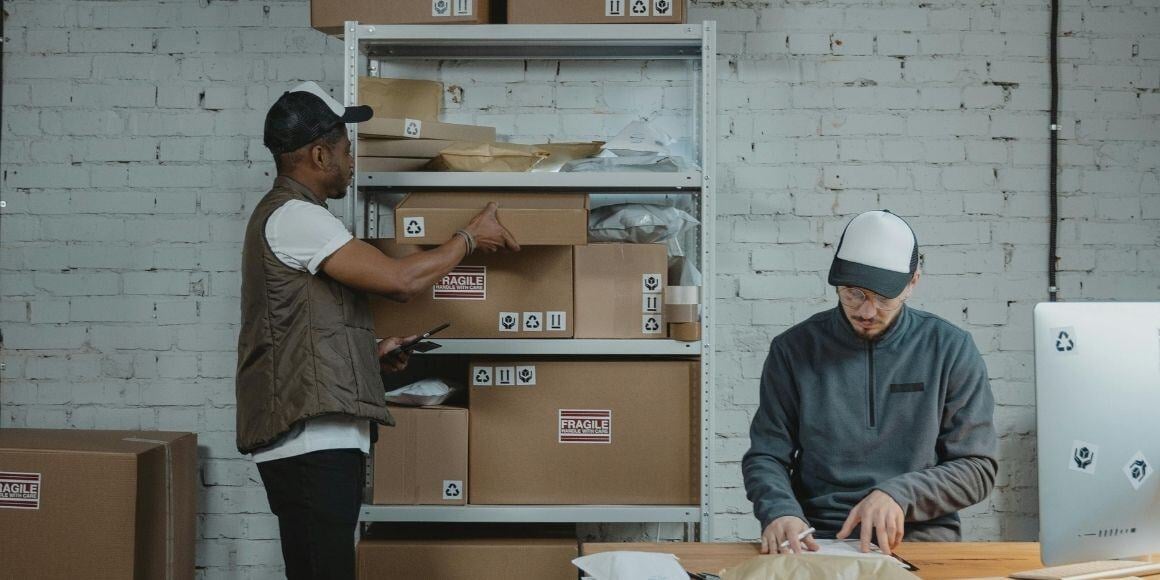 Two workers in a warehouse with a computer, collaborating on order orchestration for efficient logistics.